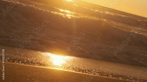Slow-motion footage of golden ocean waves gently rolling onto the sandy shore during a beautiful sunset. The warm light, shimmering reflections, and soft motion evoke a peaceful, romantic atmosphere