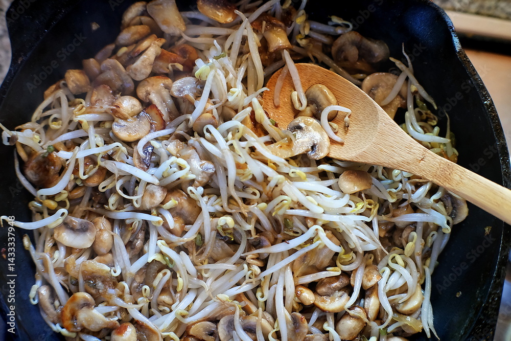 Φωτογραφία A hot pan filled with sautéed mung bean sprouts and sliced mushrooms being stirred with a wooden spoon