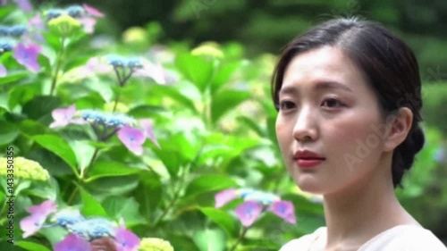 Young East Asian Woman Portrait Video in Garden. Calm beautiful female standing outdoors among green foliage and blooming hydrangea flowers in natural light.