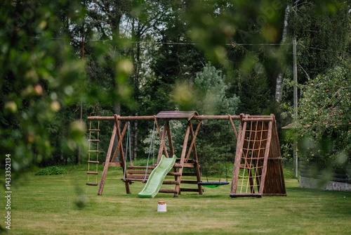 Photography Wooden outdoor playground set with a slide, swings, and climbing frames stands on a grassy lawn surrounded by trees in a peaceful natural setting