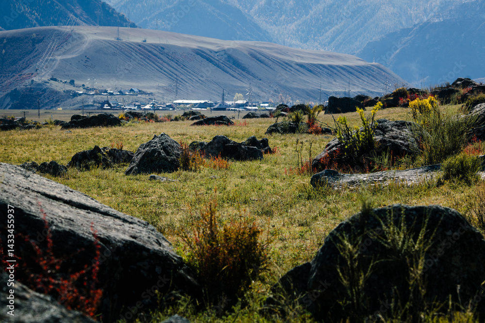A rugged landscape with boulders and mountains in the Altai Republic, with a small village visible in the background.