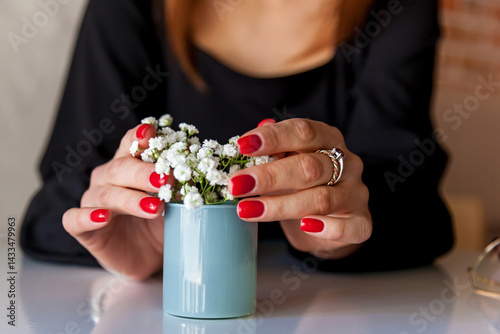 Close-up of elegant female hands with bright red manicure and silver ring gently holding a blue vase with tiny white flowers, resting on a glossy table surface