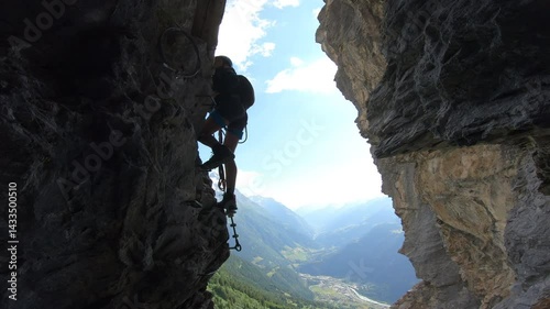 A climber emerges from a cave into daylight, silhouetted against panoramic mountain views near Silenen on the Hexensteig route.