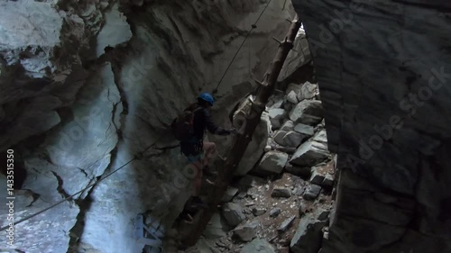 A climber balances between two steep rock faces on a wooden trunk of the Hexensteig, gripping cables as light streams above.
