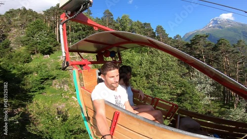 A couple enjoy the breathtaking view from a rare open-air gondola suspended over a green Swiss valley with distant alpine peaks.