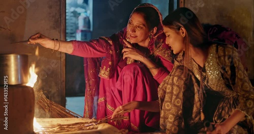 Indian villager mom with teen age female girl prepare food using earthen clay mud stove indoor house asian young adult woman lady wear red saree cooking on open fire flam inside kitchen home desi life