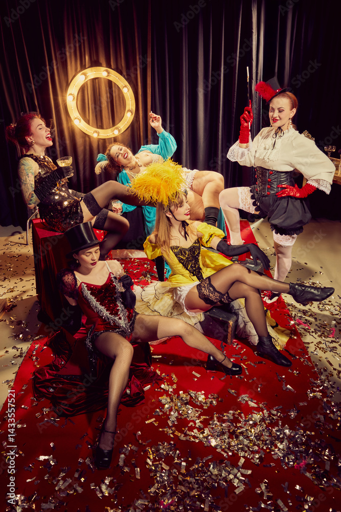 Cabaret women in vintage costumes reclining with drinks and laughter in theatrical pose on red carpet surrounded by confetti. Concept of cabaret, show, circus, retro, vintage, performance.