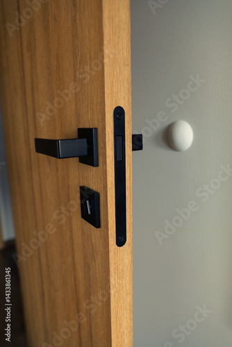 Modern wooden door with matte black handle and lock, slightly ajar, against a neutral wall with a round door stopper.