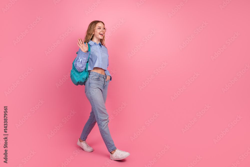 Cheerful young woman with a backpack waving hello while walking against a vibrant pink background