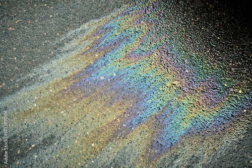 An oil slick creates a vibrant array of colors on the wet pavement, showcasing hues of blue, purple, and green. The reflections shimmer under diffused light in an urban environment.