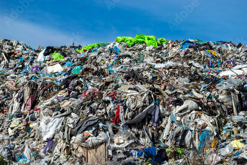 A vast accumulation of discarded clothing and fabrics fills the landfill, highlighting the issue of textile waste under bright sunlight