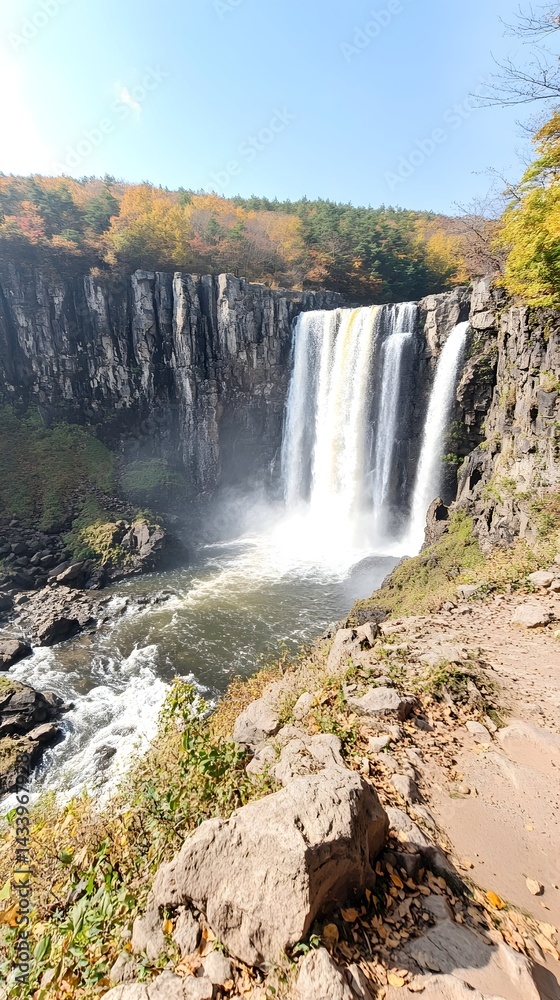 Fototapeta premium Majestic waterfall cascading down rocky cliffs, surrounded by autumnal foliage A scenic view from a rocky vantage point
