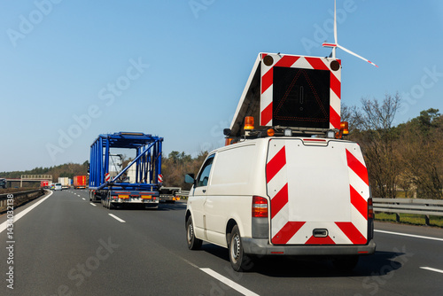 Escort van with warning sign follows a heavy-duty truck transporting large blue construction frame on multilane highway in Germany. White special cargo escort road traffic car assistance service