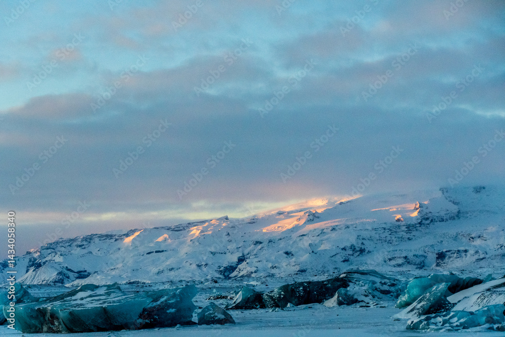 Icebergs and huge chunks for green-blueish ice fill Jokulsarlon Glacier lagoon, as the golden hour sunlight of the early morning casts a beautiful warm glow over the glacier-filled national park in