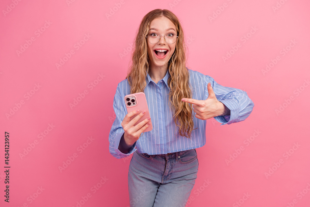 Excited young woman with wavy hair pointing to her phone, standing against a vibrant pink background while embracing a cheerful mood