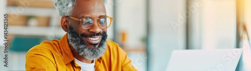 A cheerful older man with glasses smiles while working on a laptop, wearing a vibrant orange shirt in a bright, modern workspace.