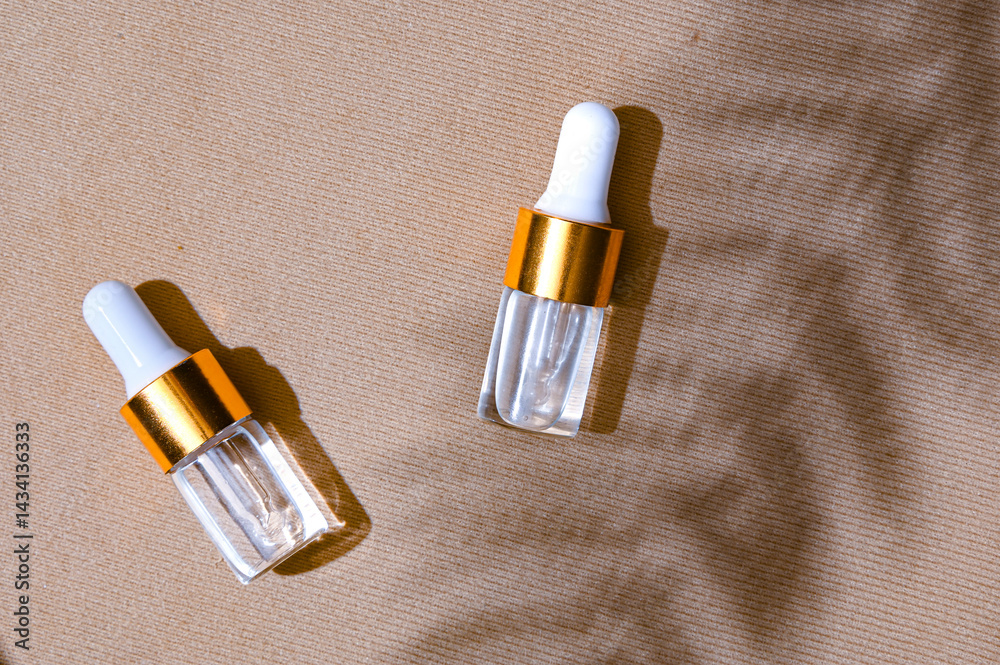 Two transparent dropper serum bottles with white caps and gold collars placed on a textured beige background with soft leaf shadows. Natural skincare branding concept with summer mood
