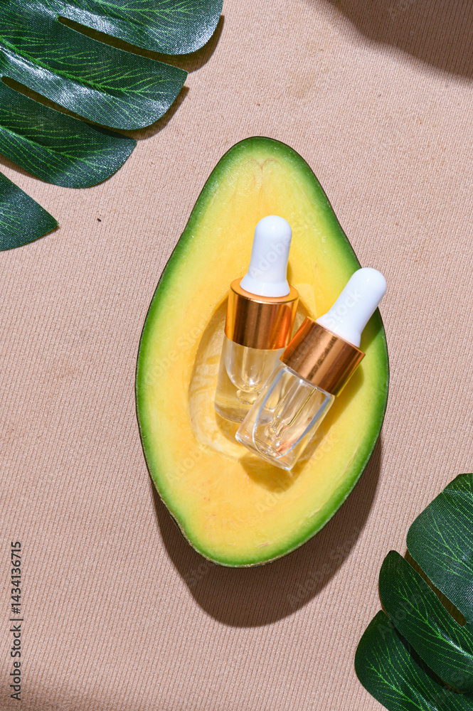 Two transparent skincare dropper bottles with golden caps placed inside a fresh avocado half, surrounded by green leaves on textured beige background. Natural beauty and organic skincare concept
