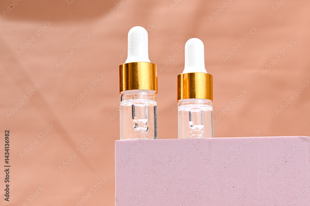 Two transparent glass serum dropper bottles with white tips and golden collars placed on a soft pink pedestal, against a peach background. Minimalist skincare branding with clean aesthetics
