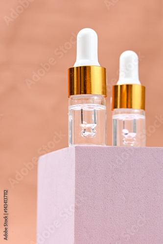 Two transparent glass serum dropper bottles with white tips and golden collars placed on a soft pink pedestal, against a peach background. Minimalist skincare branding with clean aesthetics