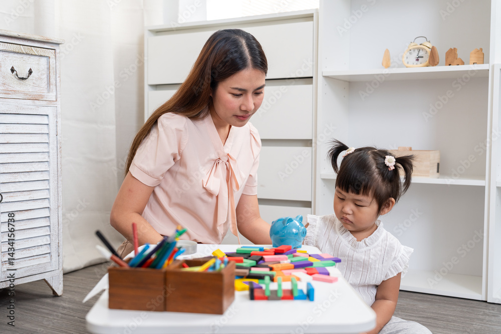Asian mother teaching preschool daughter basic saving money concept holding blue piggy bank together with colorful blocks and drawing tools on white table during playful family activity time. Asian mother teaching preschool daughter basic saving money concept holding blue piggy bank together with colorful blocks and drawing tools on white table during playful family activity time.
