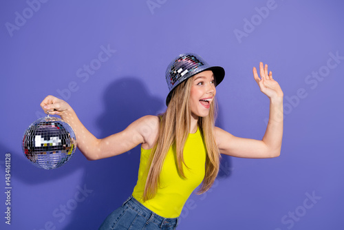 Happy young woman in vibrant casual attire holding a shiny disco ball against a purple backdrop showcasing stylish modern vibes