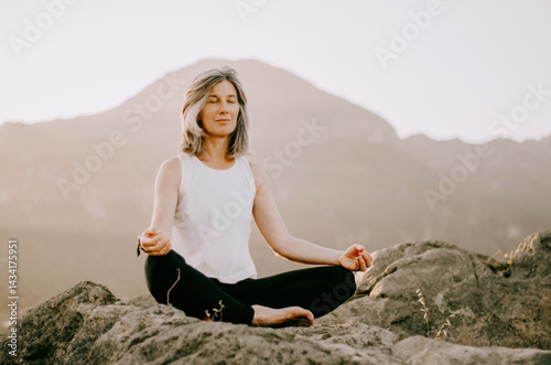 Yoga teacher meditating cross-legged on a mountain at sunset, radiating calm and wisdom