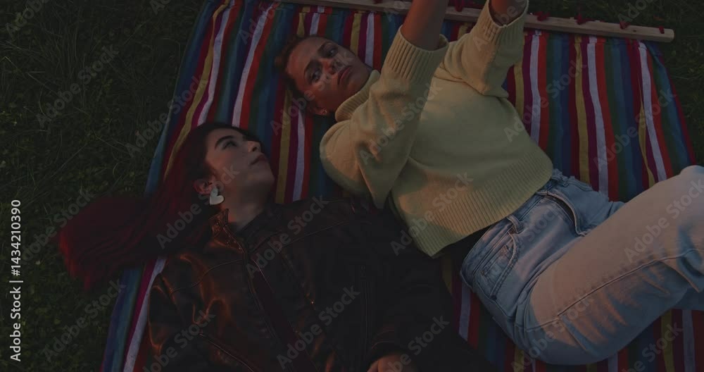 Two friends relax on a colorful blanket in a park at sunset, enjoying each other's company and taking selfies. Two friends relax on a colorful blanket in a park at sunset, enjoying each other's company and taking selfies.