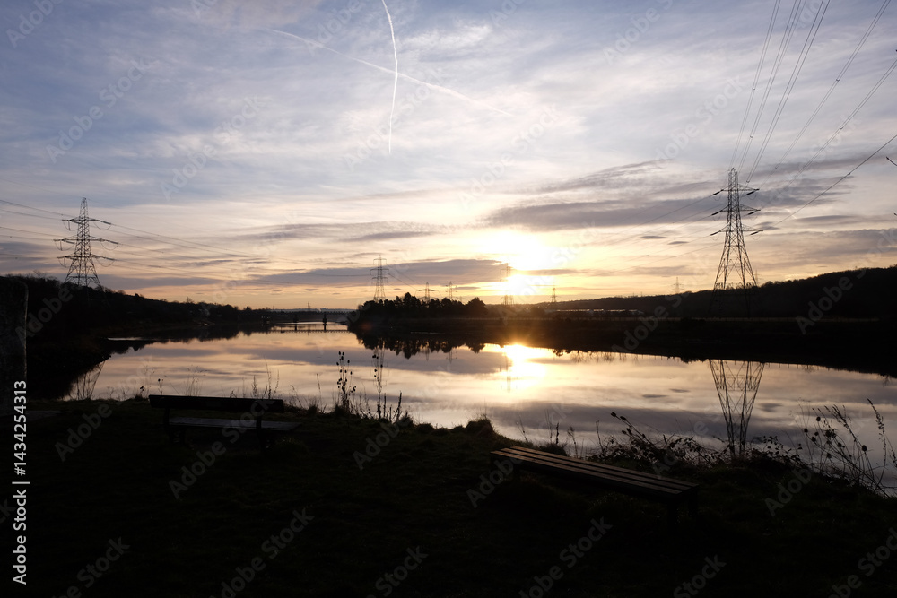 Serene sunset casts vibrant colors on the calm waters of River Tyne in Newburn, highlighting the tranquil landscape and distant power lines against the horizon.