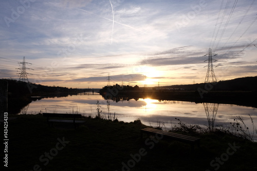 Obraz na płótnie Serene sunset casts vibrant colors on the calm waters of River Tyne in Newburn, highlighting the tranquil landscape and distant power lines against the horizon. w salonie