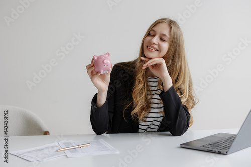 Young woman smiles while holding a pink piggy bank in a bright office setting during the day