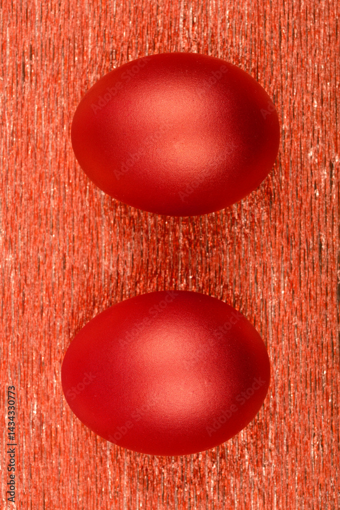 Two metallic red eggs are positioned side-by-side on a textured, shimmering red surface