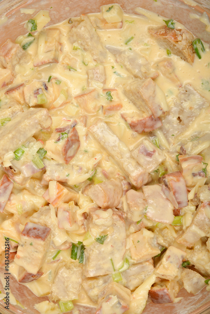 A tightly cropped top-down view shows a creamy mixture of cubed bread, vegetables, and cheese in a clear bowl, ready to be baked into a strata