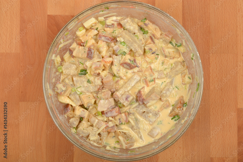 A top-down view shows a clear glass bowl filled with a creamy mixture containing cubed bread, vegetables, andcheese, prepared for a strata dish, set on a light brown wooden surface