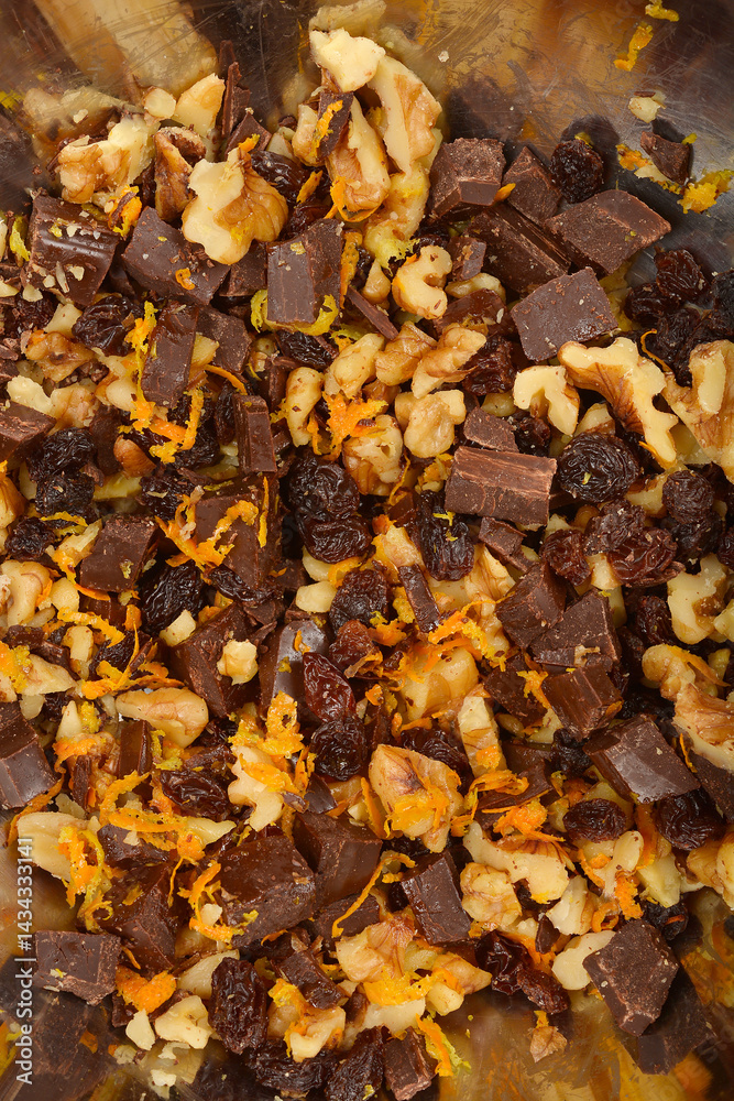 A tightly cropped top-down view shows a mixture of walnuts, dark chocolate chunks, raisins, and vibrant orange zest, possibly with some lemon zest, in a metal bowl