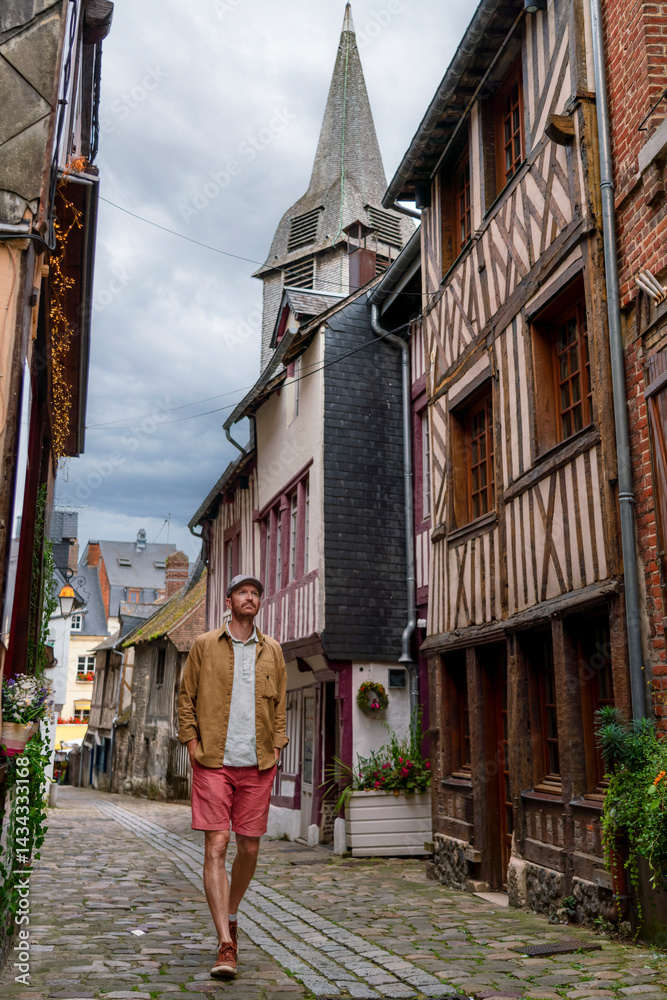 A man strolls through the cobbled, historic streets of Honfleur, a beautiful town in Normandy, surrounded by half-timbered houses and a towering church steeple under a moody sky.
