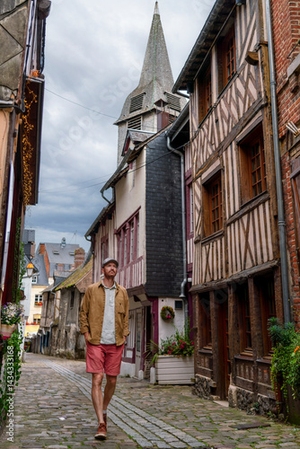 Obraz na płótnie A man strolls through the cobbled, historic streets of Honfleur, a beautiful town in Normandy, surrounded by half-timbered houses and a towering church steeple under a moody sky. w salonie