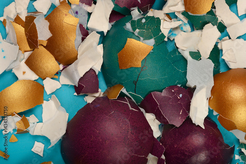 A close-up top-down view shows shattered and peeled colorful Easter eggshells in shades of gold, teal, purple, and white scattered on a light blue surface