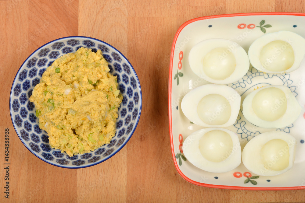 A top-down view shows a blue and white patterned bowl containing a mashed yolk, avocado, and onion mixture next to a rectangular plate with six halved hard-boiled egg whites on a wooden surface