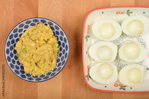Obraz na płótnie A top-down view shows a blue and white patterned bowl containing a mashed yolk, avocado, and onion mixture next to a rectangular plate with six halved hard-boiled egg whites on a wooden surface w salonie