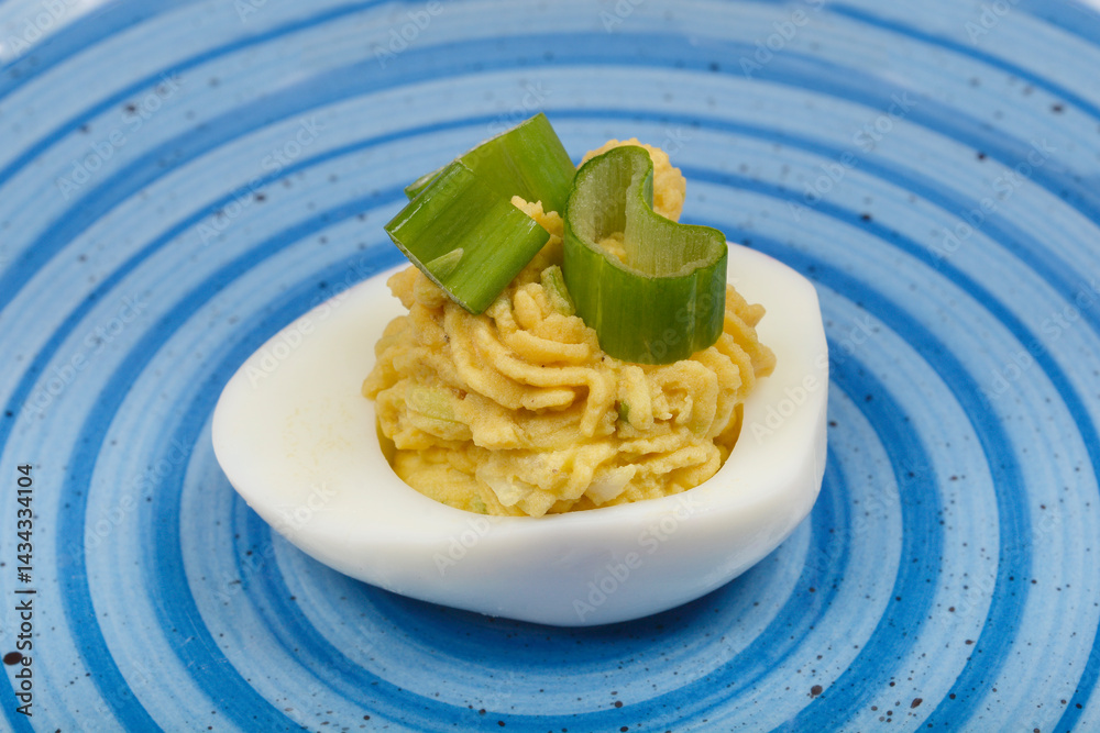 A close-up top-down view shows a deviled egg, filled with a creamy avocado and yolk mixture and garnished with green onions, on a blue and white swirled plate