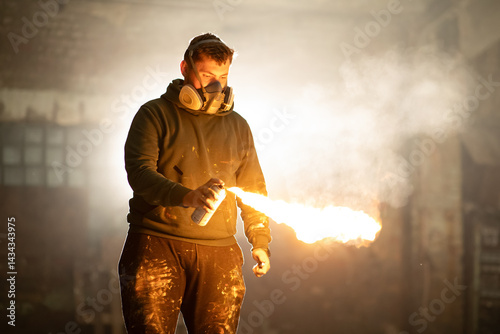 An artist wearing a mask ignites a spray can, creating a vibrant flame in an eerie, abandoned warehouse at dusk. Dust dances in the air, enhancing the dramatic effect of the scene.