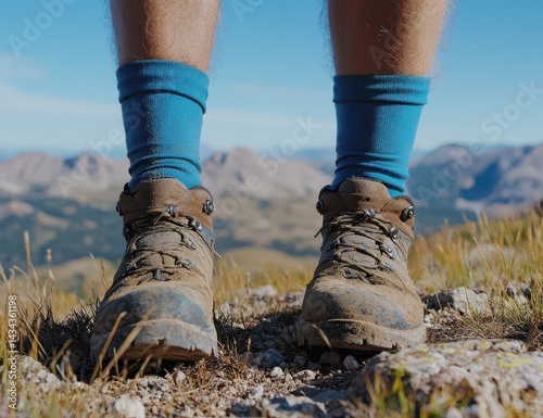 A pair of hiking boots with blue socks stand on a mountain peak, overlooking a valley with brown and green hills in the distance