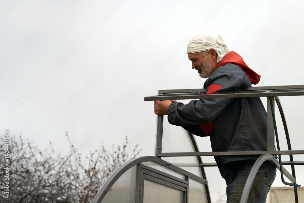 Mounting a greenhouse made of polycarbonate. An adult man attaches the end piece to the metal frame.