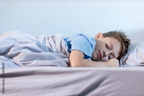 A young boy wearing a blue shirt is peacefully sleeping on his cozy bed tucked in with soft blankets and surrounded by comfortable linens.