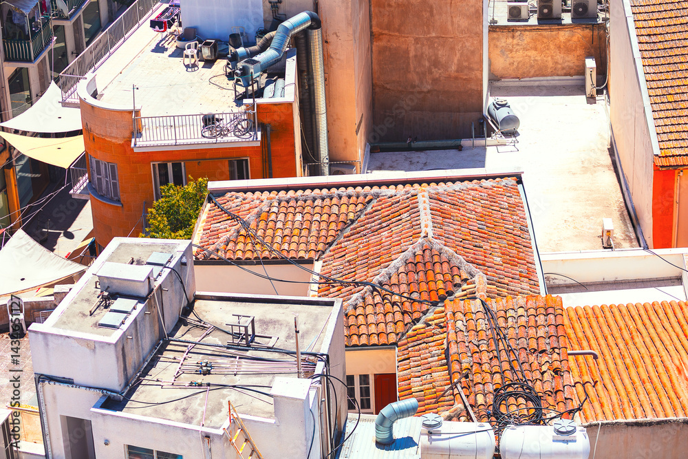 Aerial view shows a cluster of buildings with red-tiled roofs and modern rooftops, adorned with pipes, vents, and equipment. Scene includes a mix of orange and white structures under sunny sky