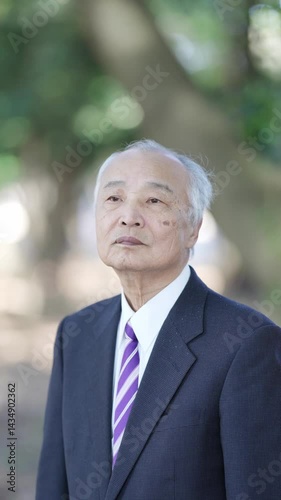 In a Japanese nature park, a Japanese man in his 70s, wearing a suit, stands and speaks calmly, surrounded by greenery and the peaceful atmosphere of nature, quietly connecting with his surroundings.