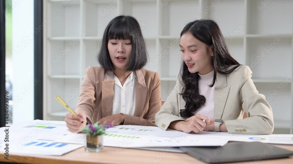 Two women are sitting at a desk, smiling and laughing while looking at a piece of paper. They seem to be discussing something important, possibly related to work or a project. The atmosphere is light Two women are sitting at a desk, smiling and laughing while looking at a piece of paper. They seem to be discussing something important, possibly related to work or a project. The atmosphere is light