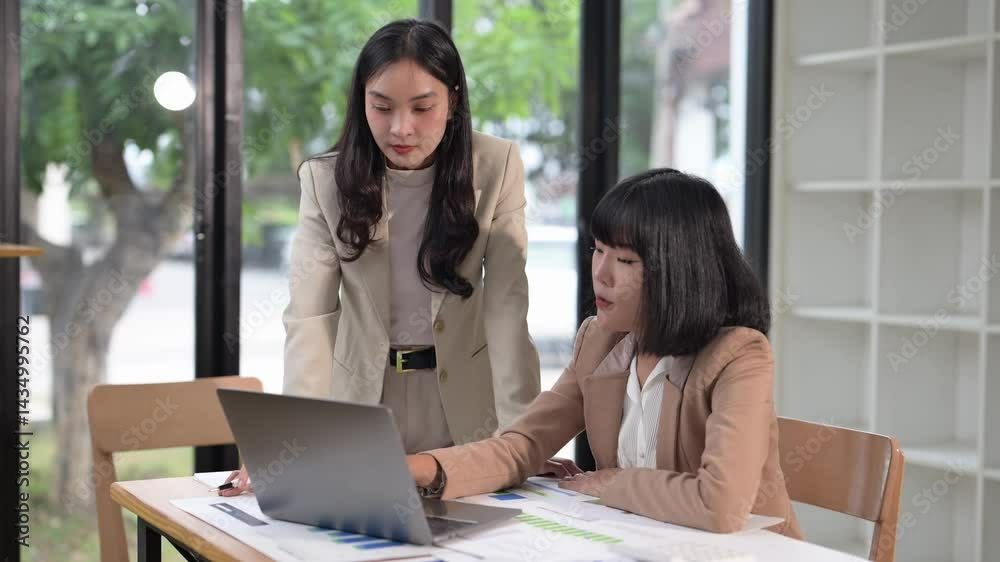 Two women are sitting at a table with a laptop in front of them. One of the women is pointing at the laptop screen Two women are sitting at a table with a laptop in front of them. One of the women is pointing at the laptop screen