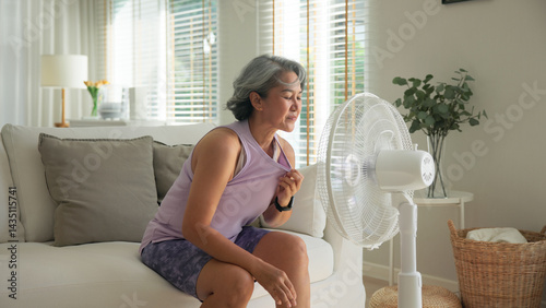 Asia adult people middle age old woman sit at sofa home feel hot flash fever use fan waving hand blowing wind face elder health care summer heat aging issue. Sad pain sweat tired stroke suffer stress.